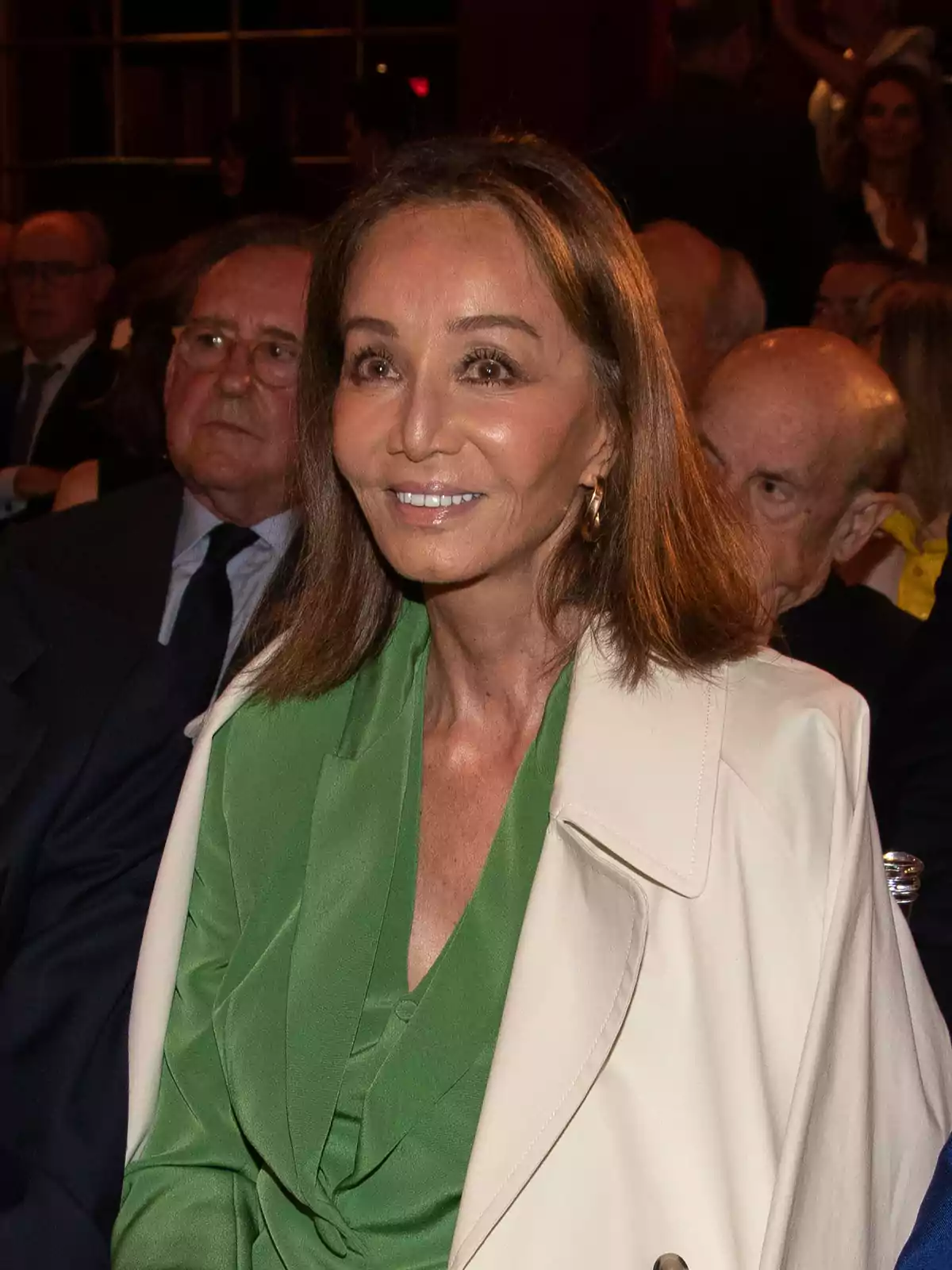 Mujer de cabello castaño y liso con chaqueta beige y blusa verde sentada en un evento rodeada de otras personas Mujer de cabello castaño y liso con chaqueta beige y blusa verde sentada en un evento rodeada de otras personas