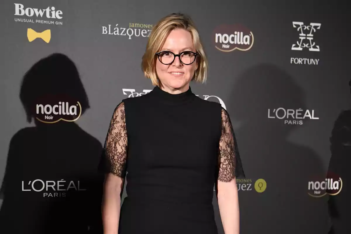 Mujer con gafas y vestido negro de encaje posando en una alfombra roja frente a un fondo con logotipos de marcas Mujer con gafas y vestido negro de encaje posando en una alfombra roja frente a un fondo con logotipos de marcas