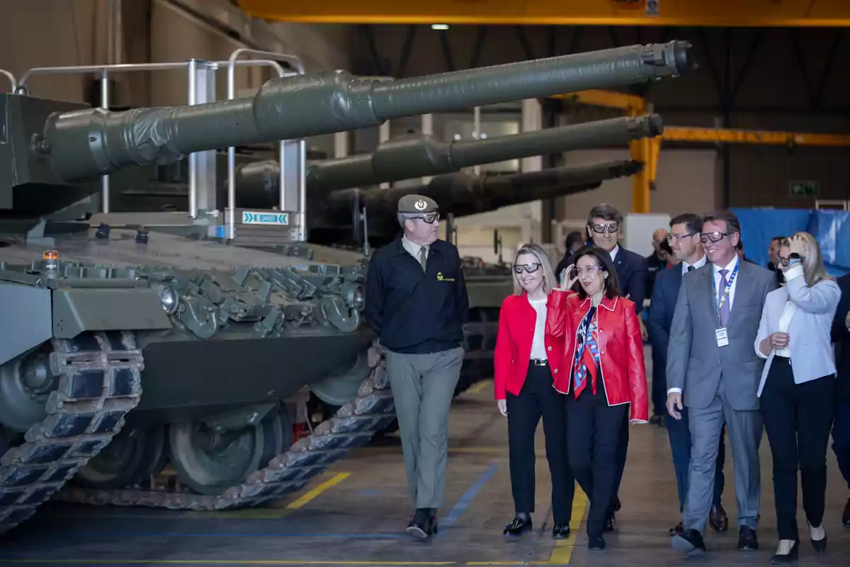 Un grupo de personas con gafas de seguridad camina junto a un tanque militar dentro de una nave industrial Un grupo de personas con gafas de seguridad camina junto a un tanque militar dentro de una nave industrial