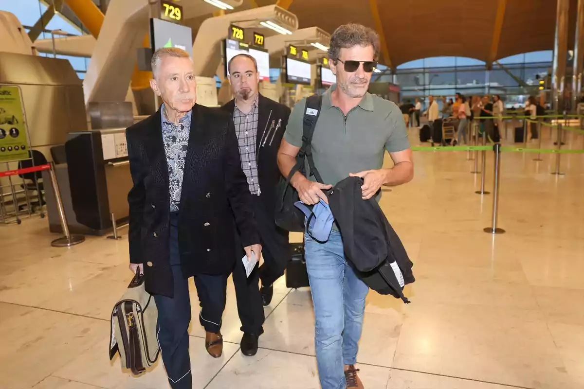 Tres hombres caminan por el área de facturación de un aeropuerto, uno de ellos lleva gafas de sol y ropa casual, otro viste un traje oscuro y lleva un maletín, mientras que el tercero los sigue de cerca Tres hombres caminan por el área de facturación de un aeropuerto, uno de ellos lleva gafas de sol y ropa casual, otro viste un traje oscuro y lleva un maletín, mientras que el tercero los sigue de cerca