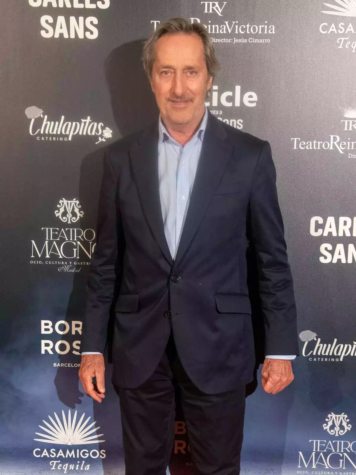 Hombre de cabello canoso y bigote, vestido con traje azul oscuro y camisa celeste, posando frente a un fondo con logotipos de marcas y eventos. Hombre de cabello canoso y bigote, vestido con traje azul oscuro y camisa celeste, posando frente a un fondo con logotipos de marcas y eventos.