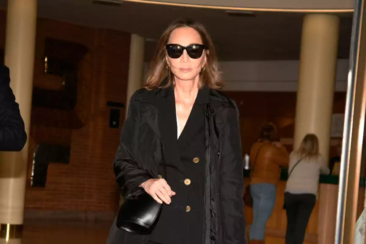 Mujer con gafas de sol y abrigo negro caminando en un interior con columnas y personas al fondo Mujer con gafas de sol y abrigo negro caminando en un interior con columnas y personas al fondo