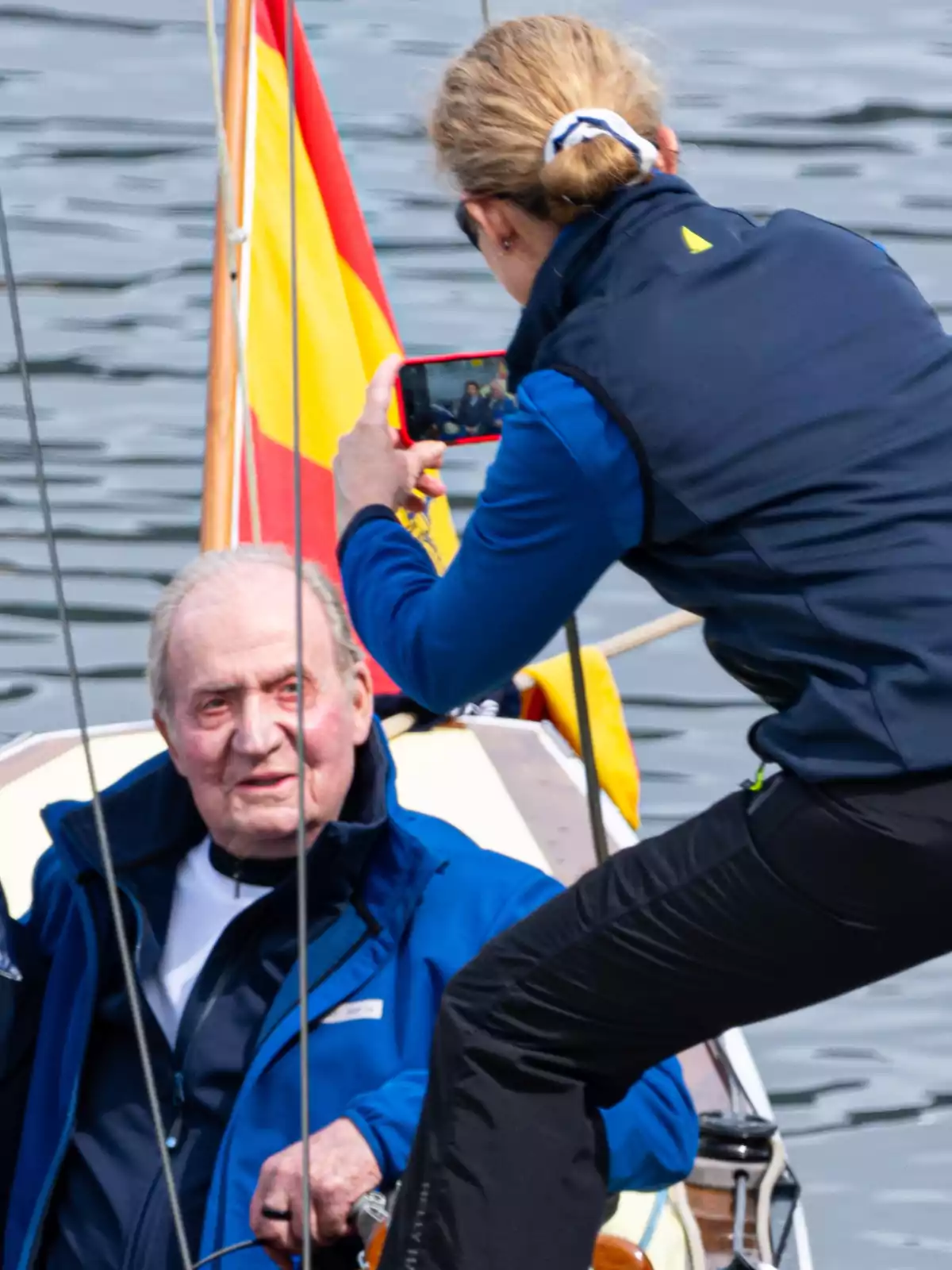 Una mujer toma una foto con su teléfono móvil a un hombre mayor que está sentado en un velero, con una bandera de España de fondo y el mar alrededor. Una mujer toma una foto con su teléfono móvil a un hombre mayor que está sentado en un velero, con una bandera de España de fondo y el mar alrededor.