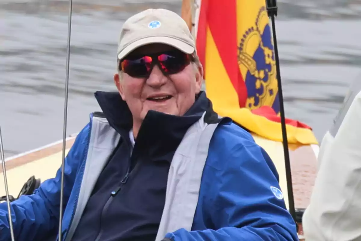 Hombre mayor sonriente con gafas de sol y gorra navegando en un barco junto a una bandera colorida al fondo Hombre mayor sonriente con gafas de sol y gorra navegando en un barco junto a una bandera colorida al fondo