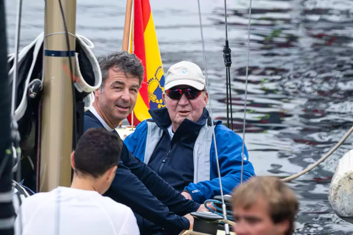 Cuatro personas navegan en un velero sobre el agua, una de ellas lleva gafas de sol y gorra, al fondo se ve una bandera de España. Cuatro personas navegan en un velero sobre el agua, una de ellas lleva gafas de sol y gorra, al fondo se ve una bandera de España.