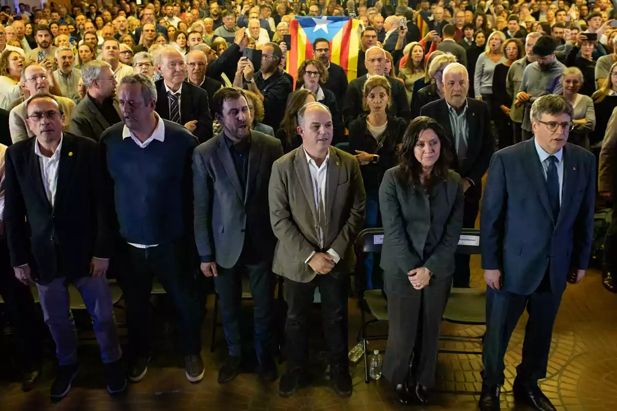 Un grupo de personas de pie en un evento multitudinario con una bandera estelada al fondo entre la multitud Un grupo de personas de pie en un evento multitudinario con una bandera estelada al fondo entre la multitud
