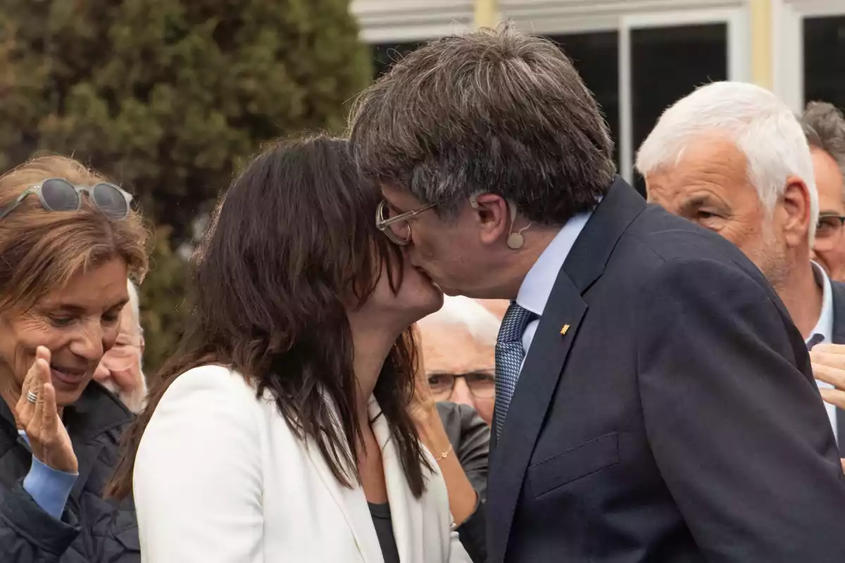 Una pareja se besa en público mientras varias personas los observan y sonríen en el fondo Una pareja se besa en público mientras varias personas los observan y sonríen en el fondo