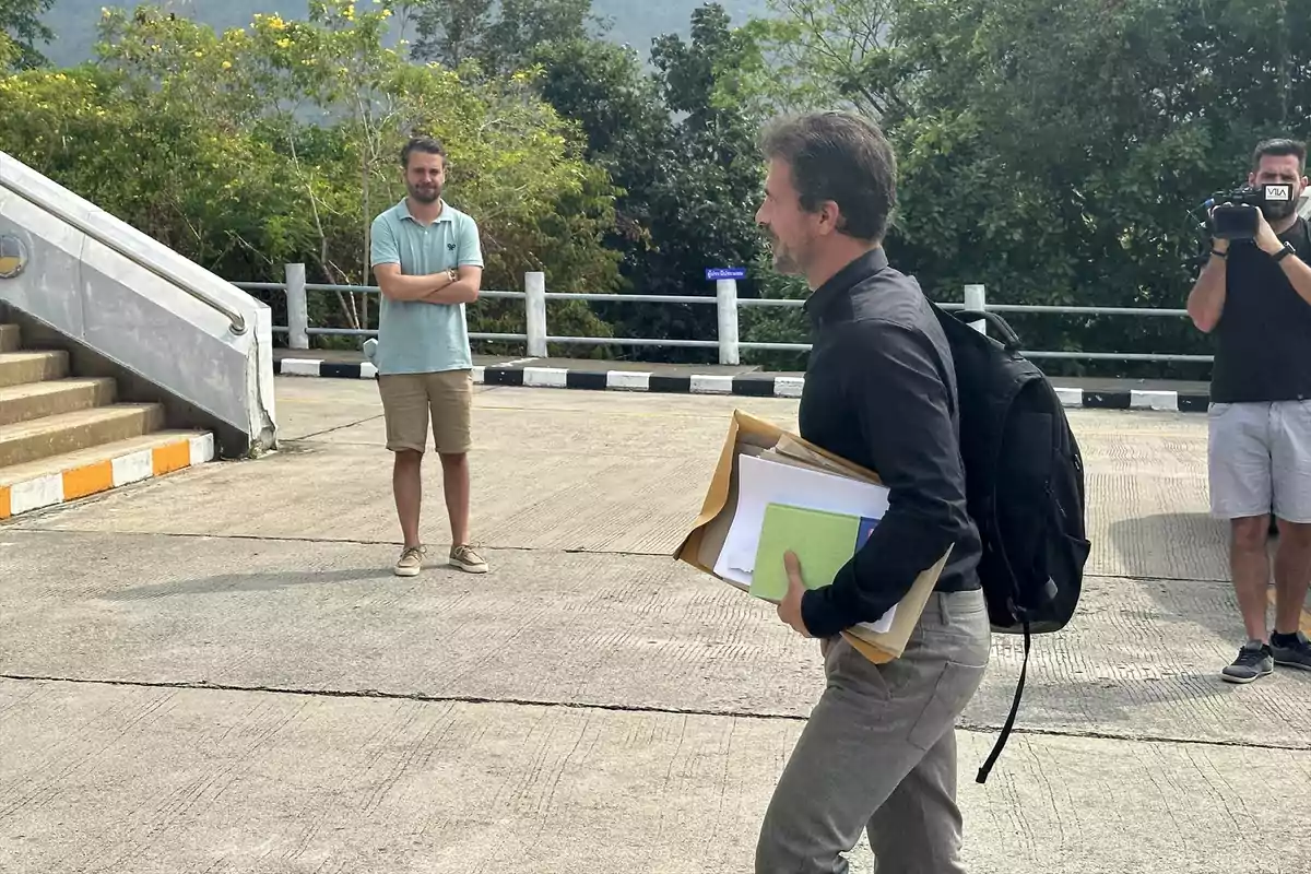 Un hombre con mochila y varios documentos camina por una acera mientras otra persona lo observa y un camarógrafo graba la escena al aire libre Un hombre con mochila y varios documentos camina por una acera mientras otra persona lo observa y un camarógrafo graba la escena al aire libre