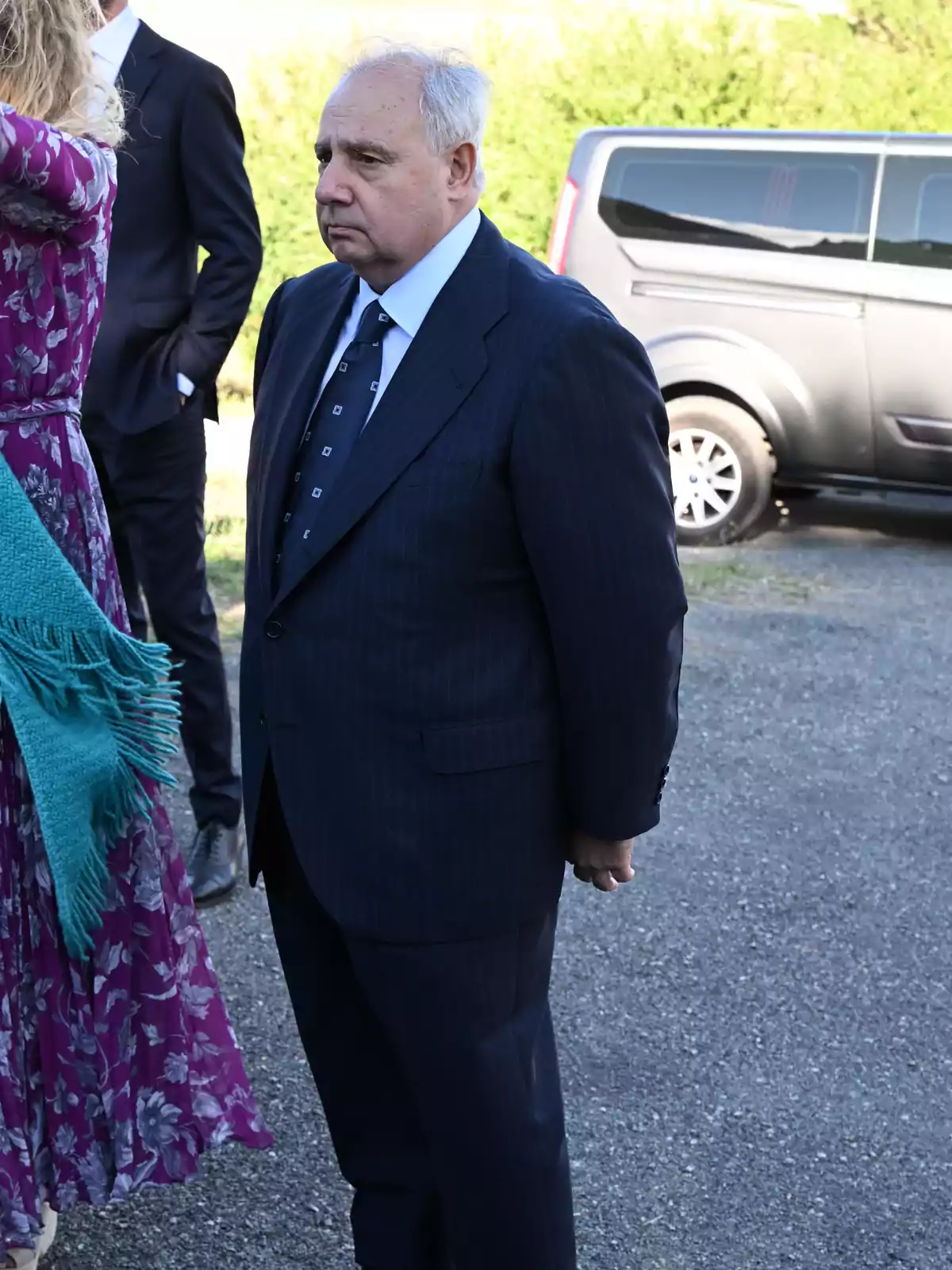 Hombre mayor de cabello canoso vistiendo un traje oscuro y corbata, de pie al aire libre junto a otras personas y una furgoneta negra estacionada al fondo Hombre mayor de cabello canoso vistiendo un traje oscuro y corbata, de pie al aire libre junto a otras personas y una furgoneta negra estacionada al fondo