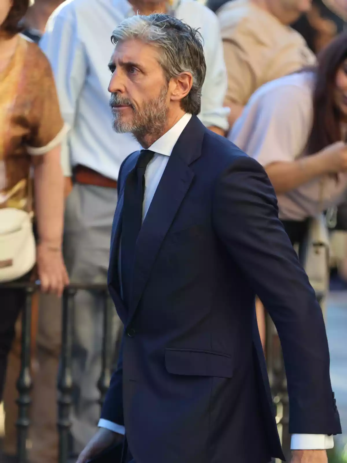Hombre de cabello canoso y barba con traje azul marino y corbata negra caminando entre una multitud en un evento formal Hombre de cabello canoso y barba con traje azul marino y corbata negra caminando entre una multitud en un evento formal