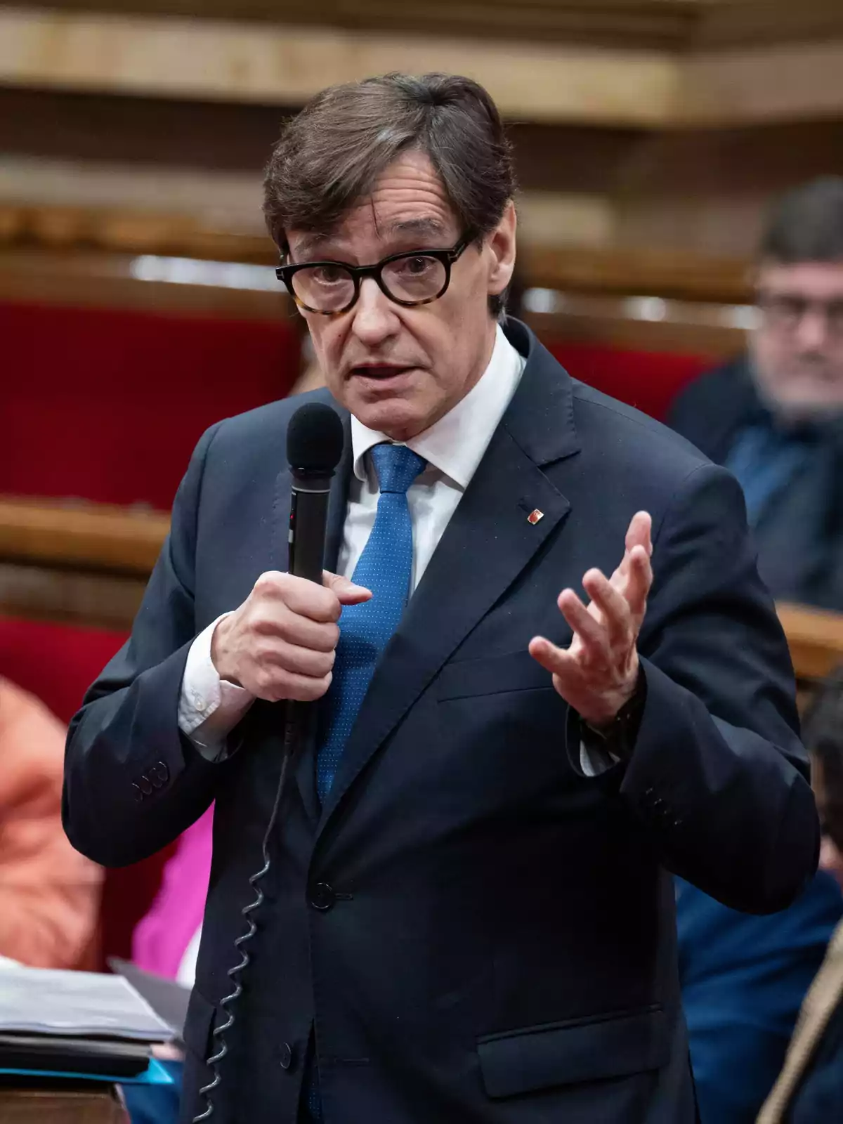 Hombre con gafas y traje azul hablando mientras sostiene un micrófono en un entorno formal Hombre con gafas y traje azul hablando mientras sostiene un micrófono en un entorno formal