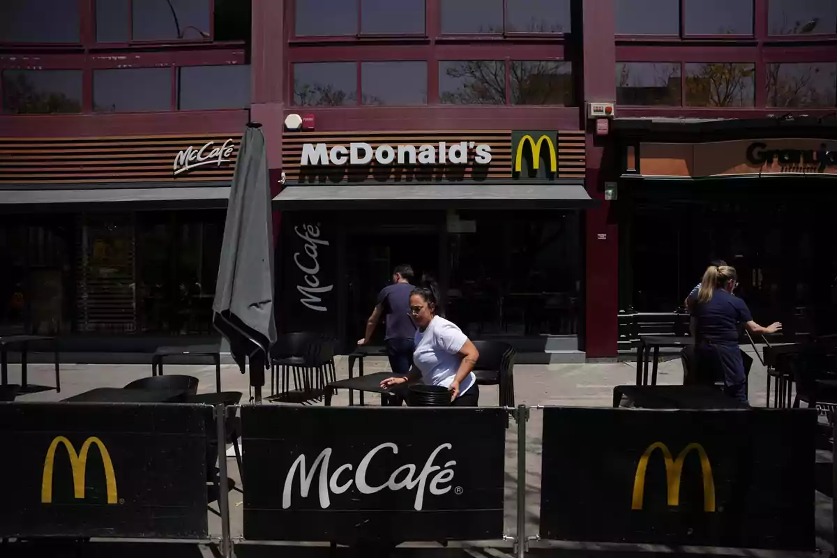 Personas acomodan mesas y sillas en la terraza de un restaurante McDonald's con letreros de McCafé al frente del local. Personas acomodan mesas y sillas en la terraza de un restaurante McDonald's con letreros de McCafé al frente del local.