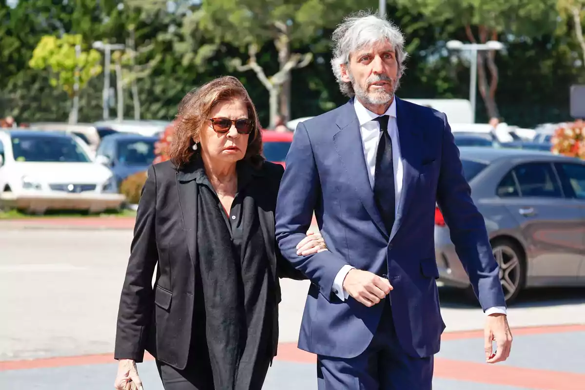 Una mujer vestida de negro y un hombre con traje azul caminan juntos al aire libre en un estacionamiento, ambos con expresión seria. Una mujer vestida de negro y un hombre con traje azul caminan juntos al aire libre en un estacionamiento, ambos con expresión seria.