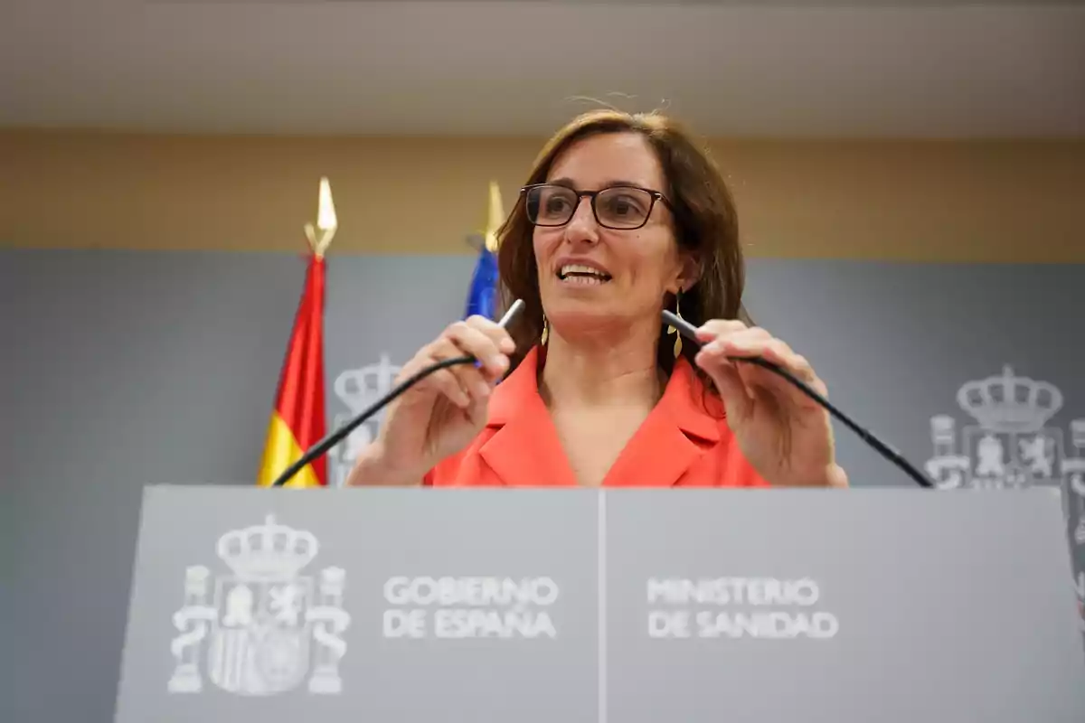 Mujer con gafas y chaqueta naranja hablando en una rueda de prensa frente a un atril del Ministerio de Sanidad del Gobierno de España con banderas al fondo Mujer con gafas y chaqueta naranja hablando en una rueda de prensa frente a un atril del Ministerio de Sanidad del Gobierno de España con banderas al fondo