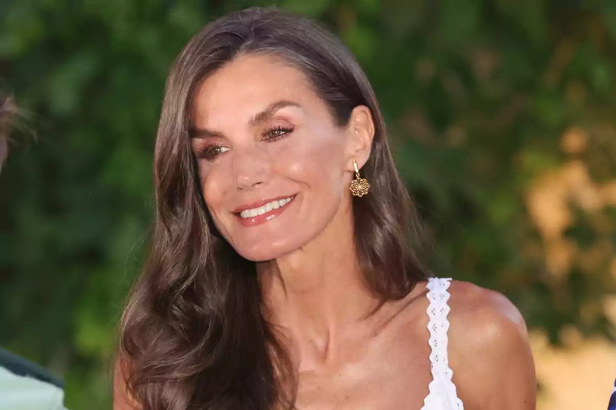 Long-haired brunette woman smiles while wearing a white dress and gold earrings with a background of blurred vegetation