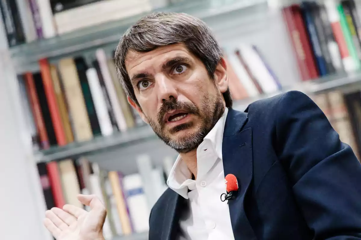 Hombre de cabello corto y barba con expresión de sorpresa o preocupación sentado frente a una estantería de libros y usando un micrófono de solapa rojo Hombre de cabello corto y barba con expresión de sorpresa o preocupación sentado frente a una estantería de libros y usando un micrófono de solapa rojo