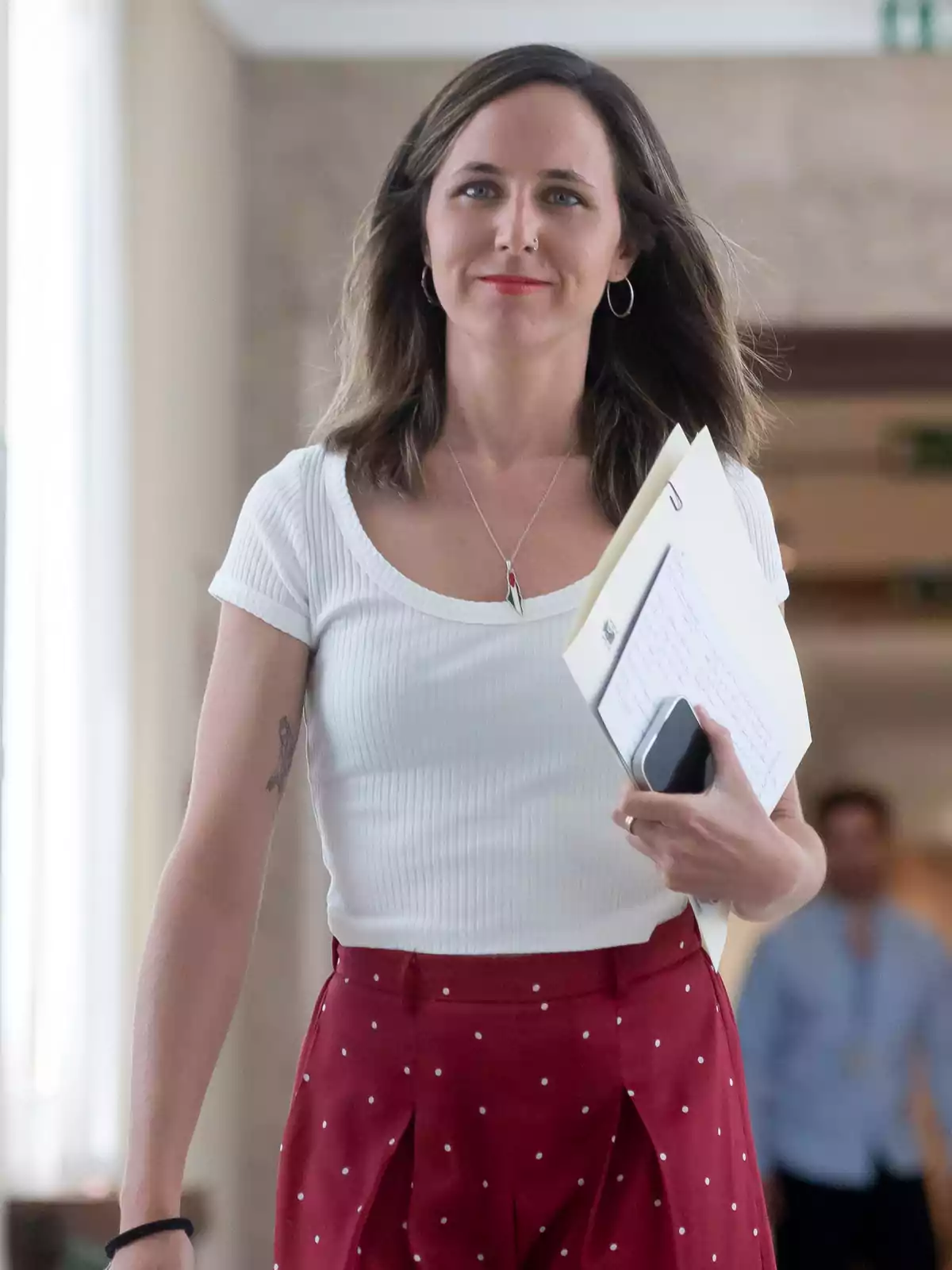 Mujer joven con blusa blanca y falda roja de lunares caminando por un pasillo mientras sostiene documentos y un teléfono móvil Mujer joven con blusa blanca y falda roja de lunares caminando por un pasillo mientras sostiene documentos y un teléfono móvil
