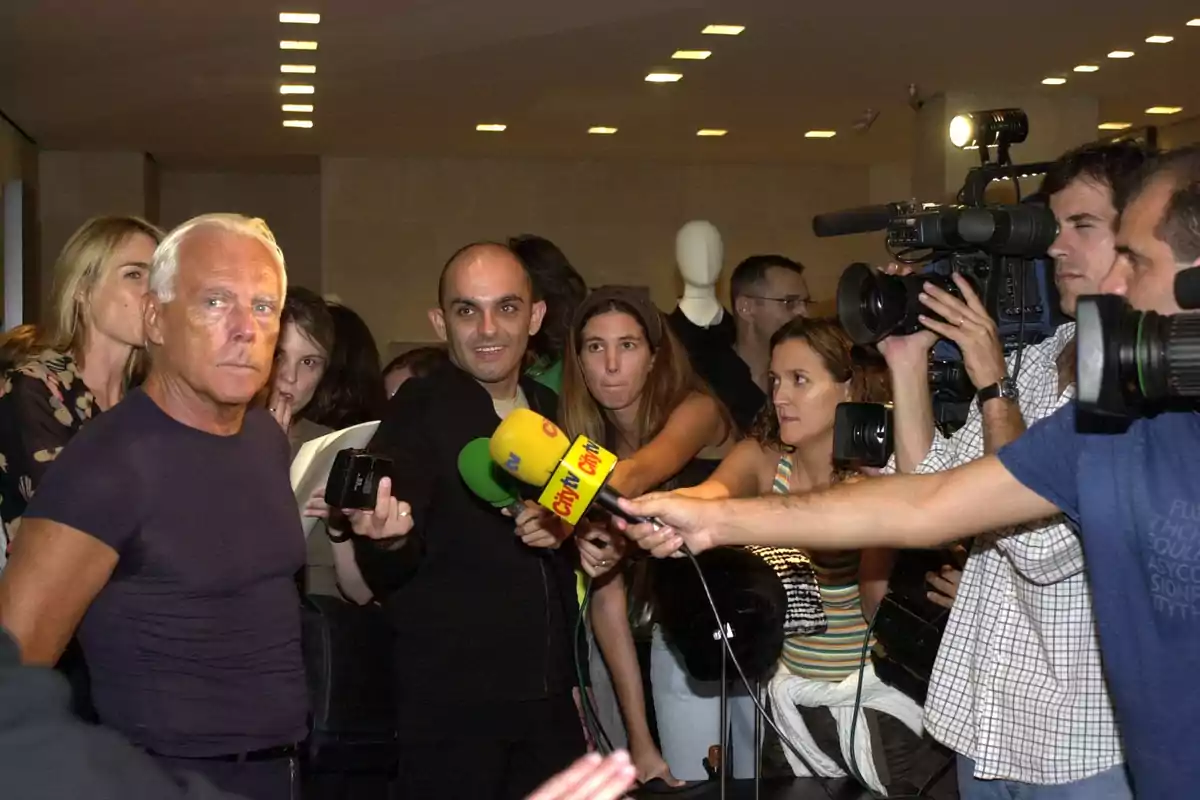 Un hombre de cabello canoso y camiseta oscura está rodeado de periodistas y camarógrafos que le apuntan con micrófonos y cámaras en un evento bajo techo Un hombre de cabello canoso y camiseta oscura está rodeado de periodistas y camarógrafos que le apuntan con micrófonos y cámaras en un evento bajo techo