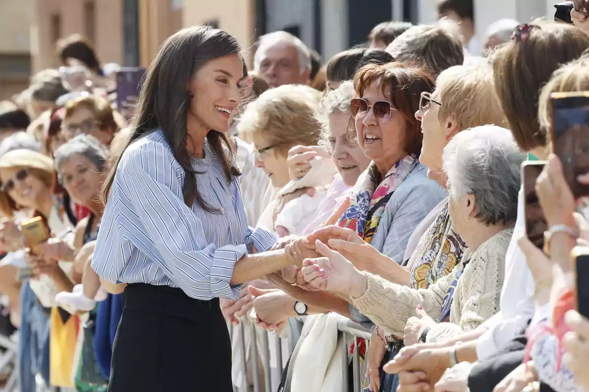A smiling woman greets and shakes hands with several elderly people standing behind a fence at an outdoor event. A smiling woman greets and shakes hands with several elderly people standing behind a fence at an outdoor event.