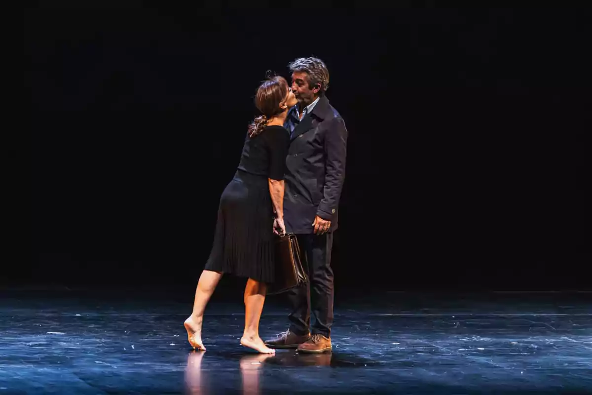 Una mujer descalza y un hombre elegantemente vestido se encuentran en el escenario oscuro mientras ella lo besa en la mejilla y sostiene un maletín Una mujer descalza y un hombre elegantemente vestido se encuentran en el escenario oscuro mientras ella lo besa en la mejilla y sostiene un maletín