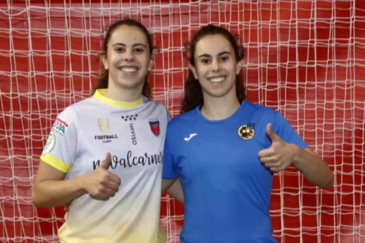 Dos jóvenes deportistas sonríen y posan con el pulgar hacia arriba frente a una portería de fútbol sala, una lleva camiseta blanca y la otra azul.