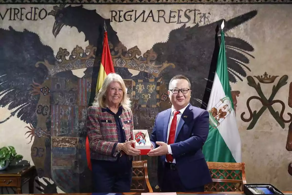 Dos personas sonríen mientras sostienen un obsequio en una sala decorada con un mural de un águila y banderas oficiales al fondo Dos personas sonríen mientras sostienen un obsequio en una sala decorada con un mural de un águila y banderas oficiales al fondo