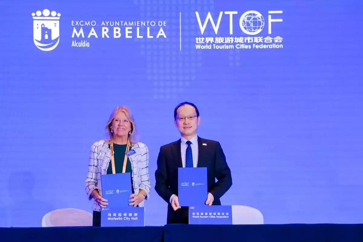Dos personas de pie sosteniendo carpetas azules frente a un fondo azul con los logotipos del Ayuntamiento de Marbella y la World Tourism Cities Federation