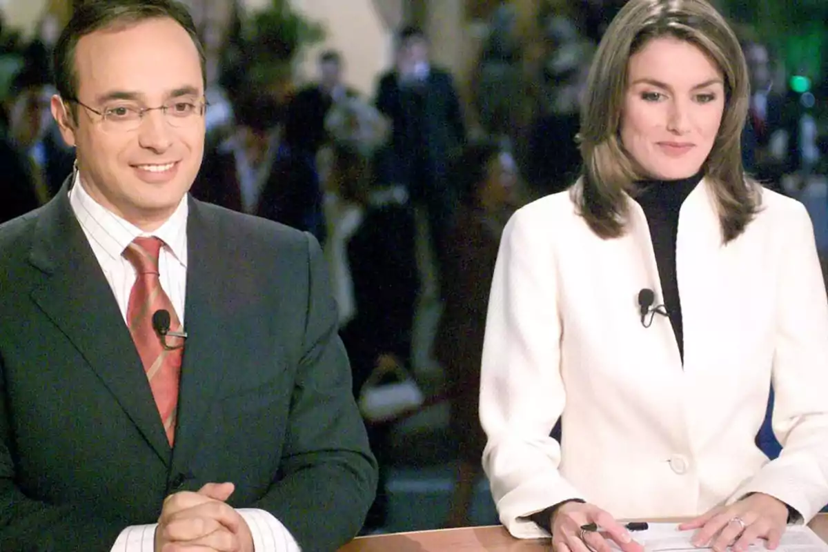 Two news anchors are sitting at a desk, both wearing lapel microphones and dressed formally—the man in a dark suit and red tie, the woman in a white jacket and black sweater—as they look ahead in a television studio. Two news anchors are sitting at a desk, both wearing lapel microphones and dressed formally—the man in a dark suit and red tie, the woman in a white jacket and black sweater—as they look ahead in a television studio.