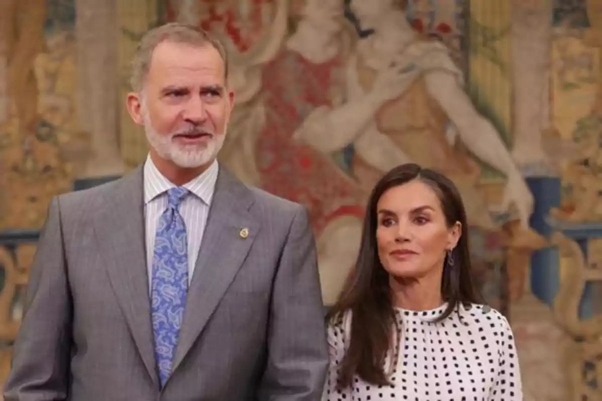 A bearded man in a gray suit stands next to a woman with long, loose hair wearing a white blouse with black polka dots as they pose in front of a decorative tapestry. A bearded man in a gray suit stands next to a woman with long, loose hair wearing a white blouse with black polka dots as they pose in front of a decorative tapestry.