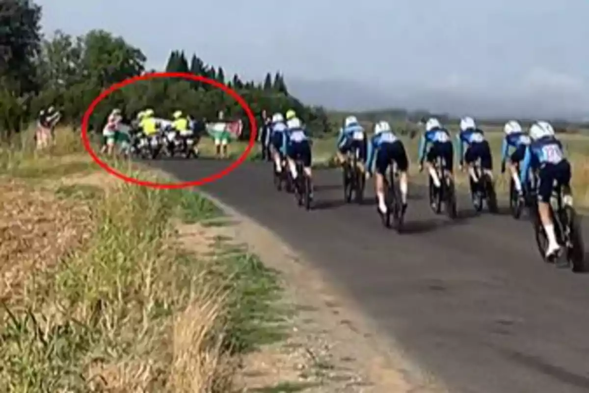 Grupo de ciclistas compitiendo en una carretera mientras al fondo se observa un accidente marcado con un círculo rojo Grupo de ciclistas compitiendo en una carretera mientras al fondo se observa un accidente marcado con un círculo rojo