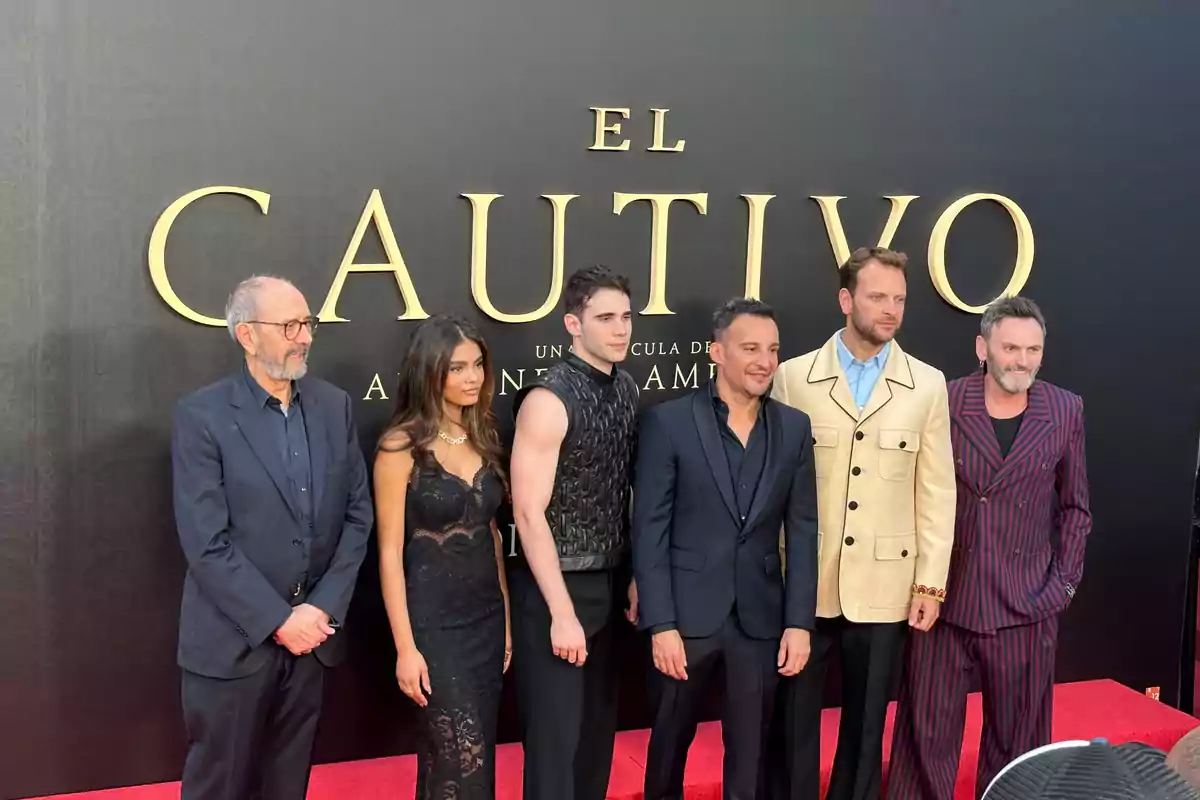 Grupo de seis personas posando en una alfombra roja frente a un fondo negro con letras doradas que dicen El Cautivo Grupo de seis personas posando en una alfombra roja frente a un fondo negro con letras doradas que dicen El Cautivo