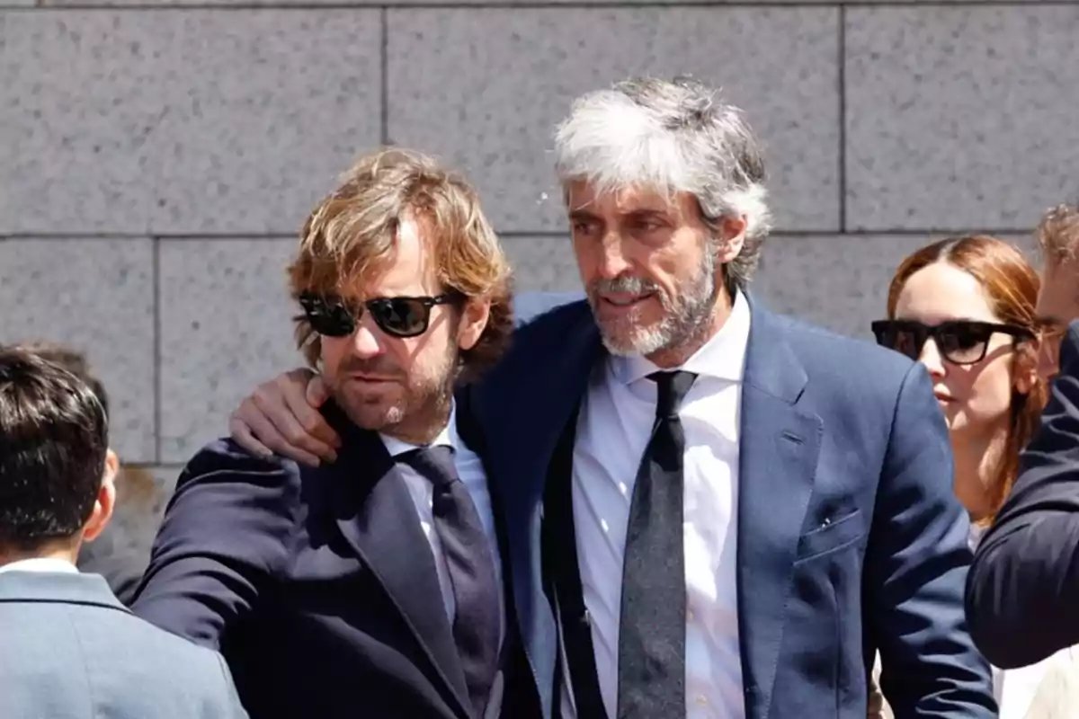 Dos hombres vestidos con traje oscuro y corbata, uno de ellos con el brazo sobre el hombro del otro, ambos llevan gafas de sol y están al aire libre junto a otras personas Dos hombres vestidos con traje oscuro y corbata, uno de ellos con el brazo sobre el hombro del otro, ambos llevan gafas de sol y están al aire libre junto a otras personas