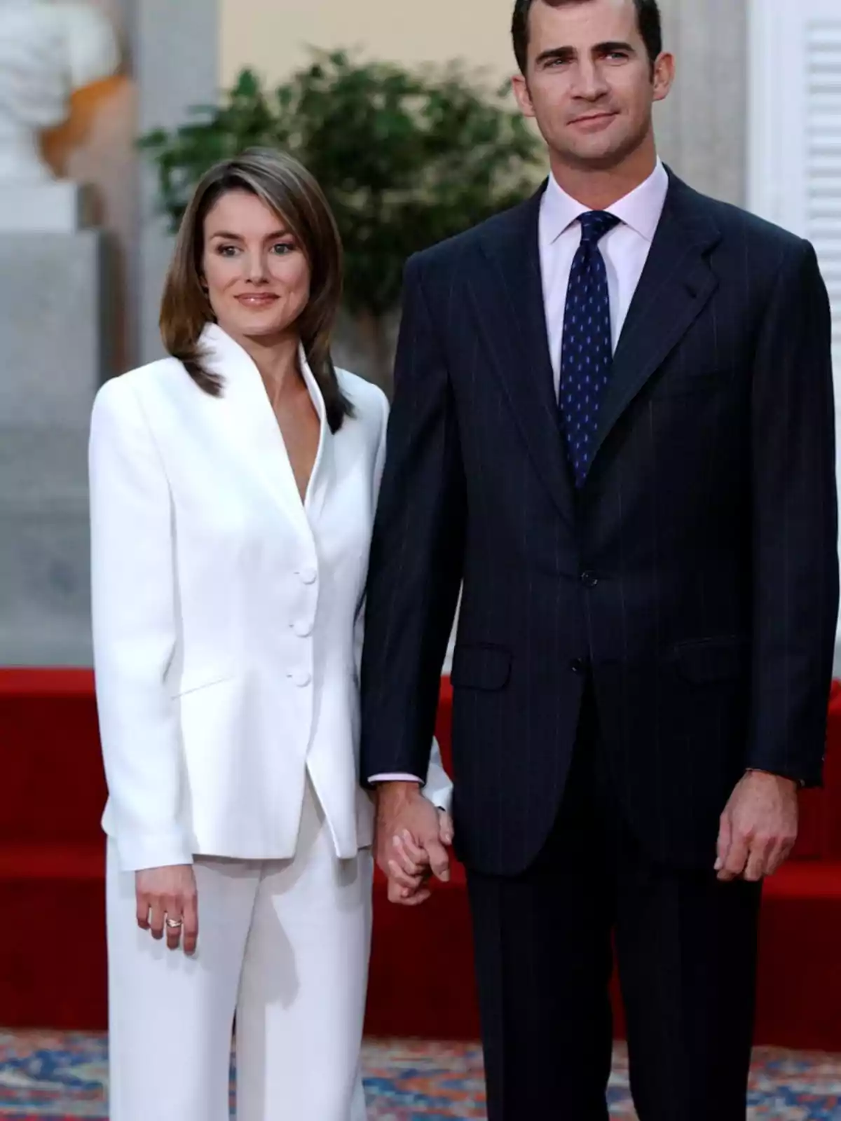 Una pareja elegantemente vestida posa de la mano en un entorno formal con alfombra y fondo de plantas Una pareja elegantemente vestida posa de la mano en un entorno formal con alfombra y fondo de plantas