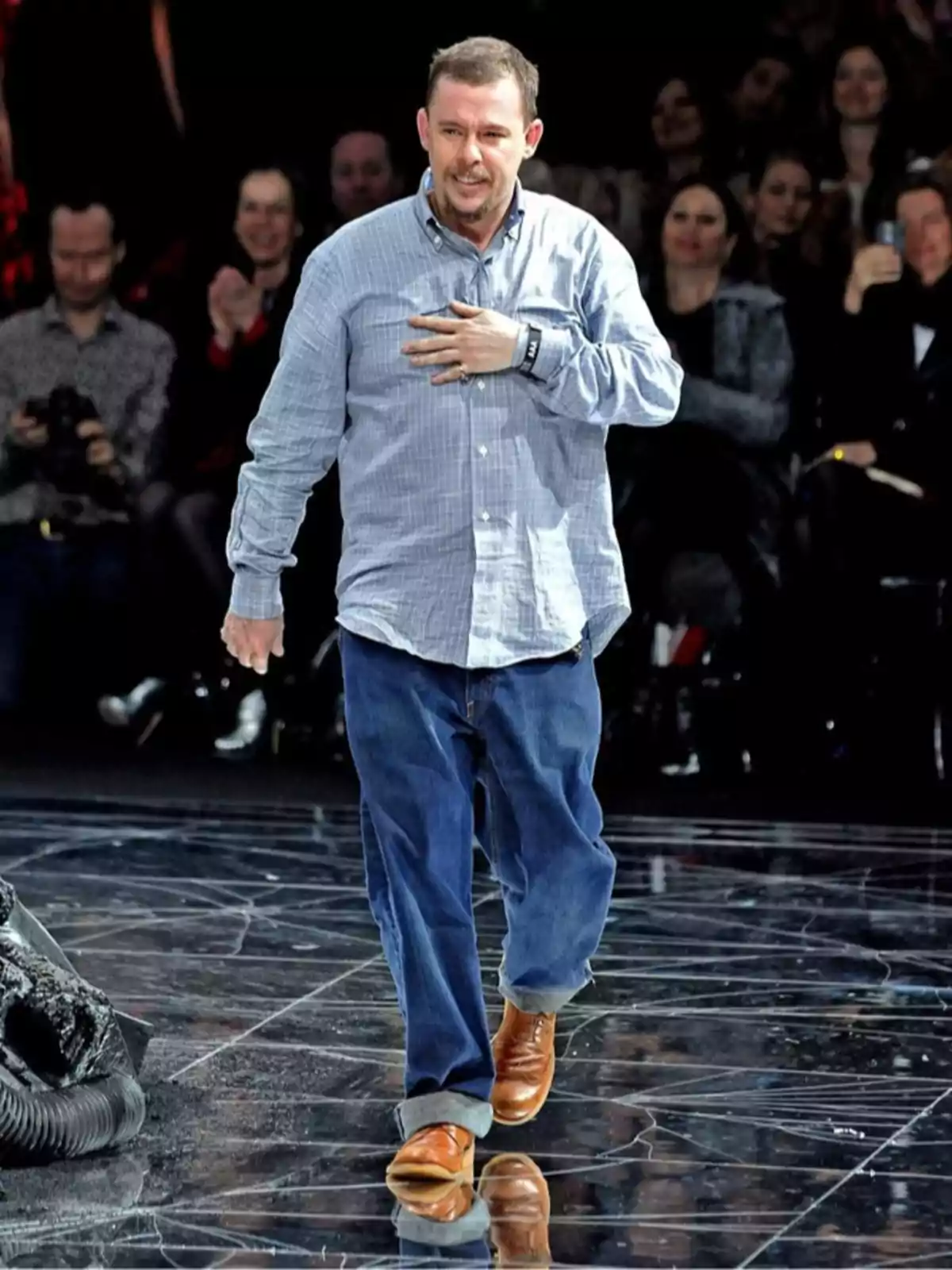 Hombre con camisa de cuadros y pantalón de mezclilla caminando sobre una pasarela mientras el público lo observa y aplaude Hombre con camisa de cuadros y pantalón de mezclilla caminando sobre una pasarela mientras el público lo observa y aplaude