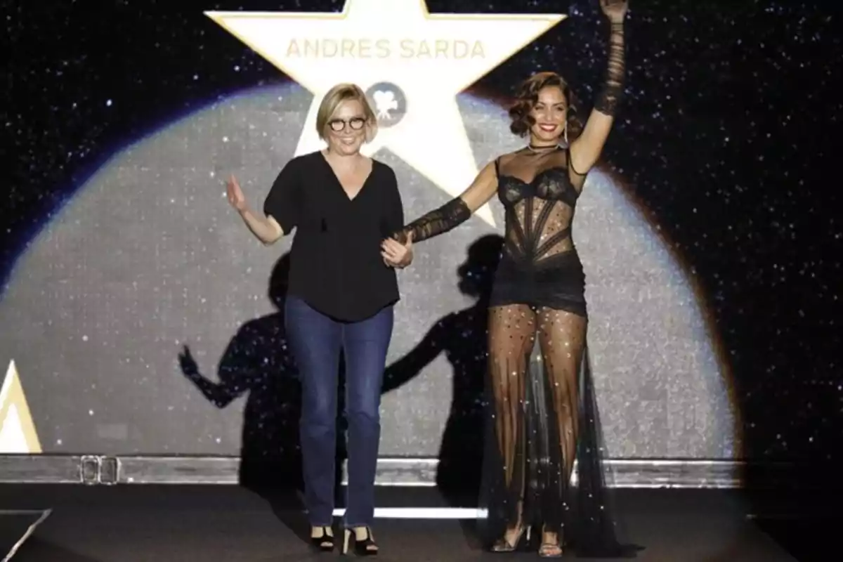 Dos mujeres caminan tomadas de la mano sobre una pasarela, una de ellas lleva un conjunto negro transparente y la otra viste ropa casual, al fondo se ve una estrella con el nombre Andres Sarda. Dos mujeres caminan tomadas de la mano sobre una pasarela, una de ellas lleva un conjunto negro transparente y la otra viste ropa casual, al fondo se ve una estrella con el nombre Andres Sarda.