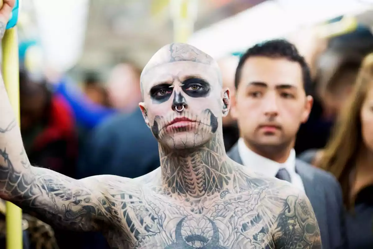 Hombre calvo con tatuajes de calavera en la cara y el cuerpo viajando en transporte público rodeado de otras personas Hombre calvo con tatuajes de calavera en la cara y el cuerpo viajando en transporte público rodeado de otras personas