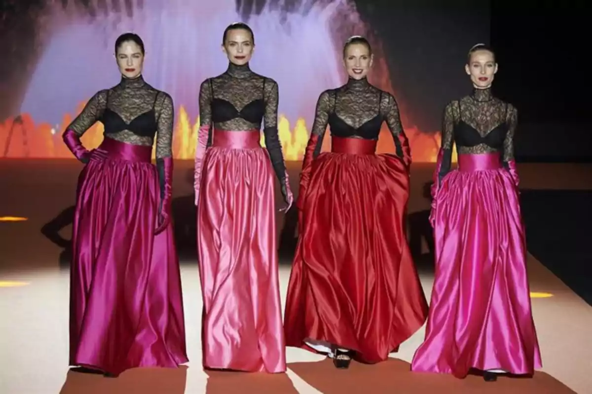 Cuatro modelos desfilan en una pasarela usando faldas largas de satén en tonos rosa y rojo con blusas de encaje negro y guantes largos a juego, con un fondo iluminado que simula una fuente de agua. Cuatro modelos desfilan en una pasarela usando faldas largas de satén en tonos rosa y rojo con blusas de encaje negro y guantes largos a juego, con un fondo iluminado que simula una fuente de agua.