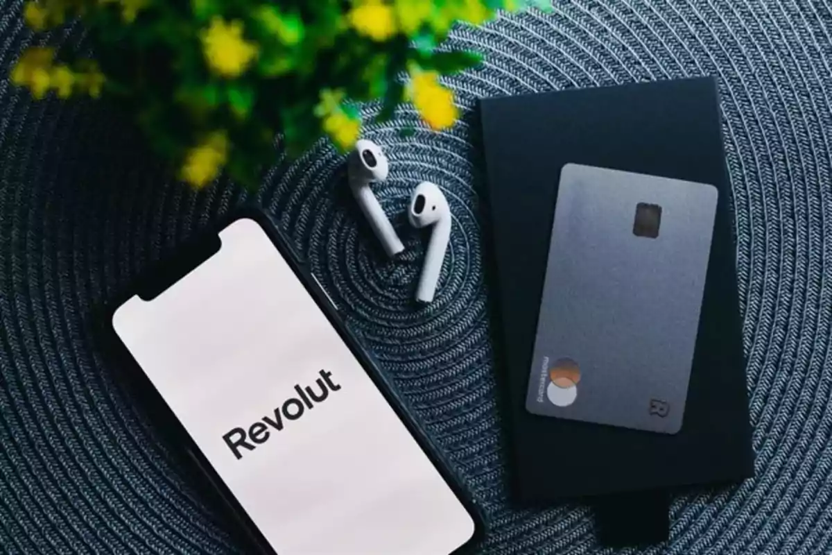 Teléfono móvil con la pantalla encendida mostrando la palabra Revolut junto a unos audífonos inalámbricos y una tarjeta bancaria sobre una superficie tejida con una planta en la esquina superior izquierda Teléfono móvil con la pantalla encendida mostrando la palabra Revolut junto a unos audífonos inalámbricos y una tarjeta bancaria sobre una superficie tejida con una planta en la esquina superior izquierda