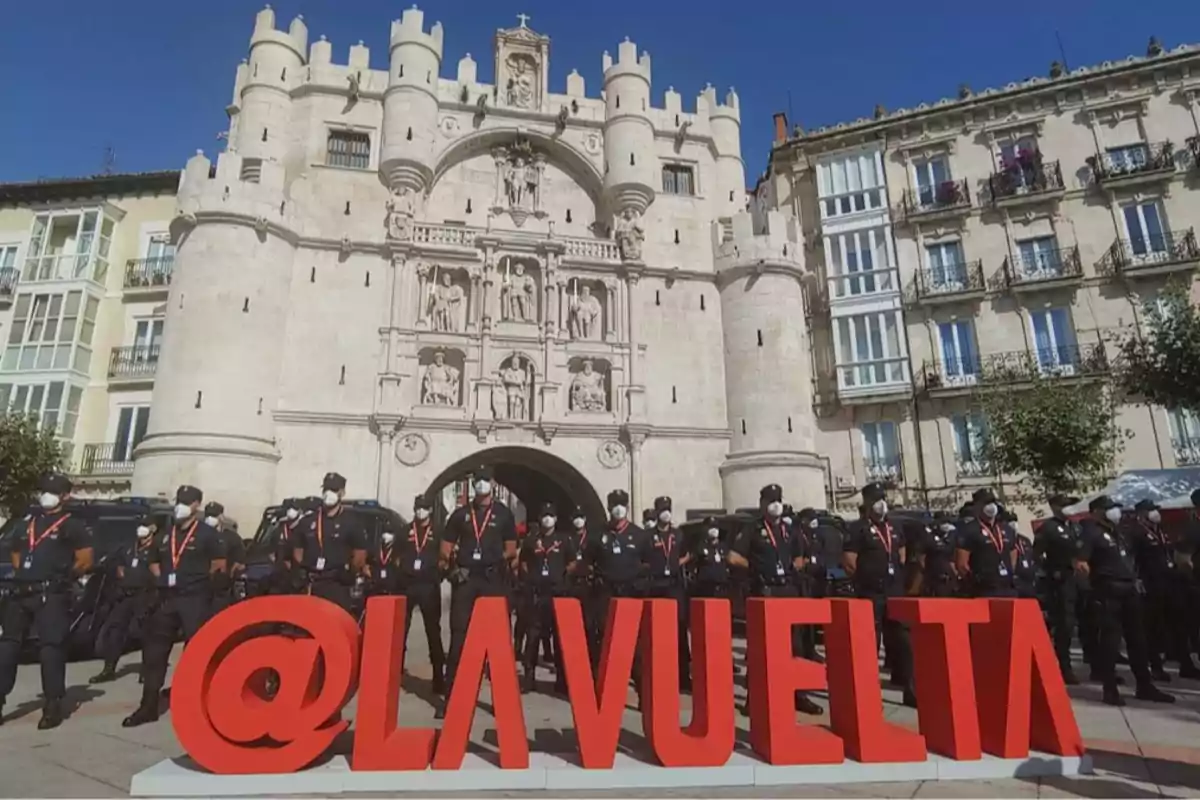 Un grupo de policías con mascarillas está de pie detrás de un letrero rojo de gran tamaño que dice LAVUELTA frente a un edificio histórico de piedra con torres y esculturas en una plaza urbana Un grupo de policías con mascarillas está de pie detrás de un letrero rojo de gran tamaño que dice LAVUELTA frente a un edificio histórico de piedra con torres y esculturas en una plaza urbana