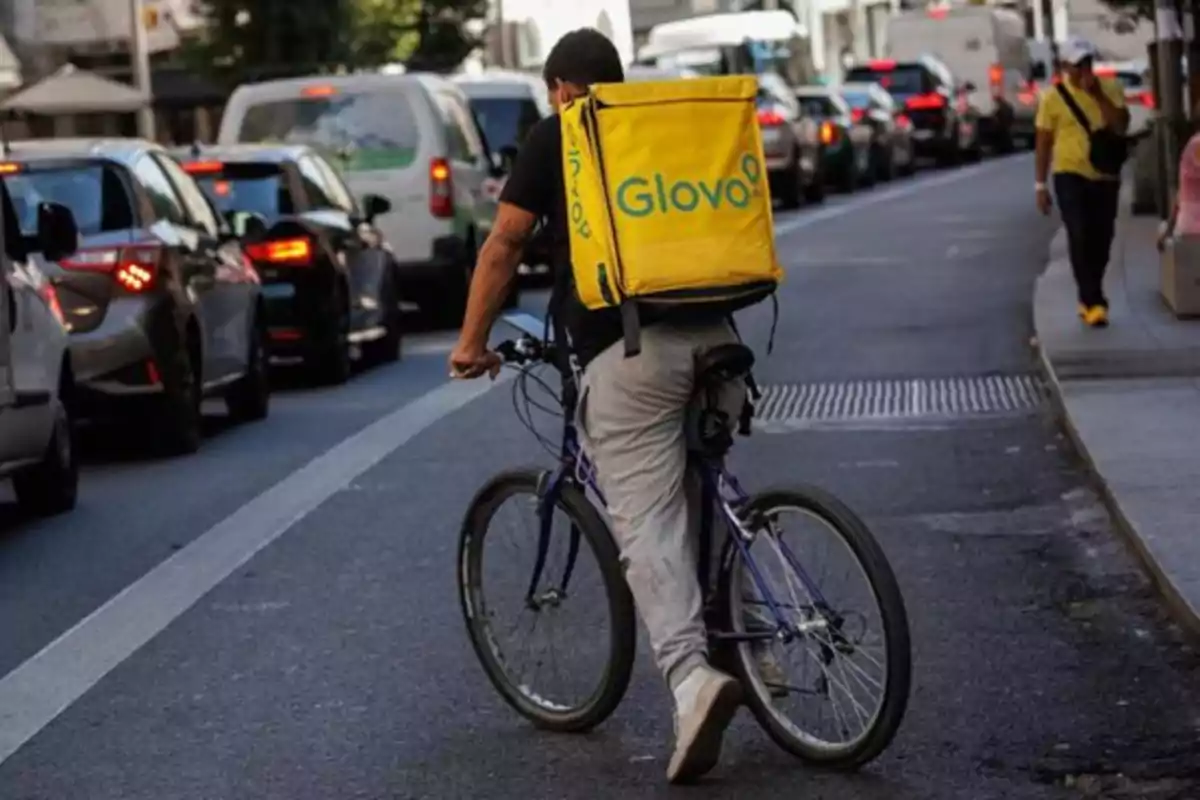Repartidor de Glovo caminando junto a su bicicleta en una calle con tráfico de autos Repartidor de Glovo caminando junto a su bicicleta en una calle con tráfico de autos