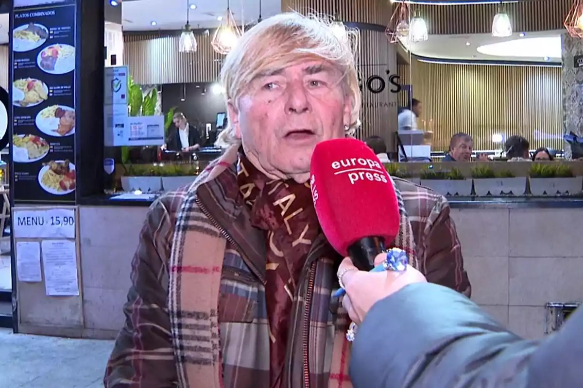 Un hombre mayor con cabello rubio y ropa de cuadros es entrevistado en la calle frente a un restaurante mientras sostiene un micrófono rojo de Europa Press Un hombre mayor con cabello rubio y ropa de cuadros es entrevistado en la calle frente a un restaurante mientras sostiene un micrófono rojo de Europa Press
