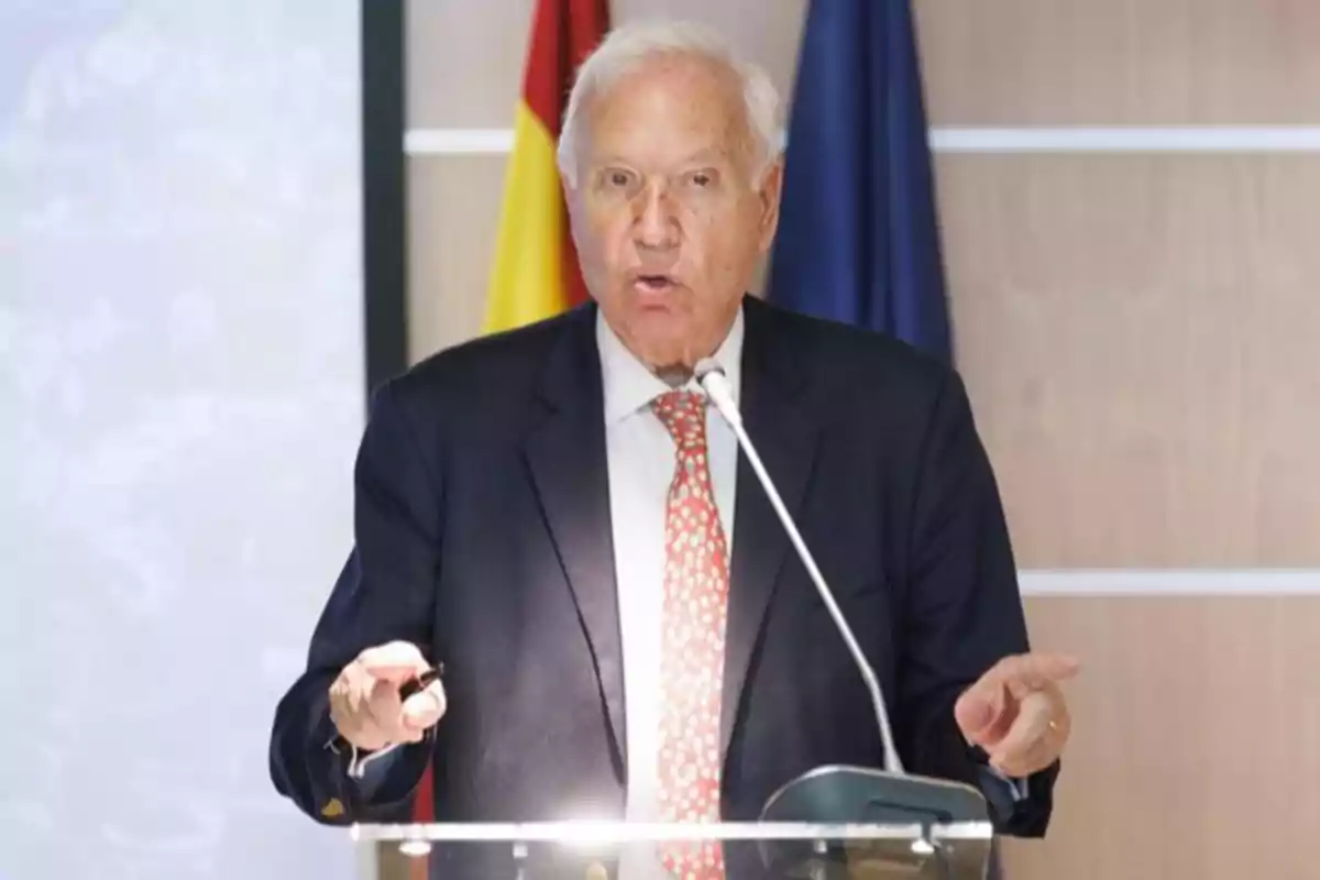 Hombre mayor de cabello canoso hablando en un podio con micrófono, vistiendo traje oscuro y corbata roja con fondo de banderas. Hombre mayor de cabello canoso hablando en un podio con micrófono, vistiendo traje oscuro y corbata roja con fondo de banderas.