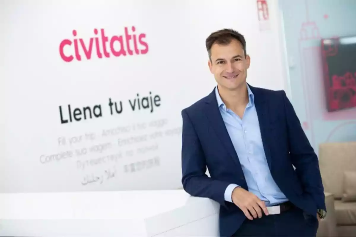 Hombre con traje azul sonriendo y posando junto a una pared con el logo de Civitatis y el eslogan Llena tu viaje Hombre con traje azul sonriendo y posando junto a una pared con el logo de Civitatis y el eslogan Llena tu viaje
