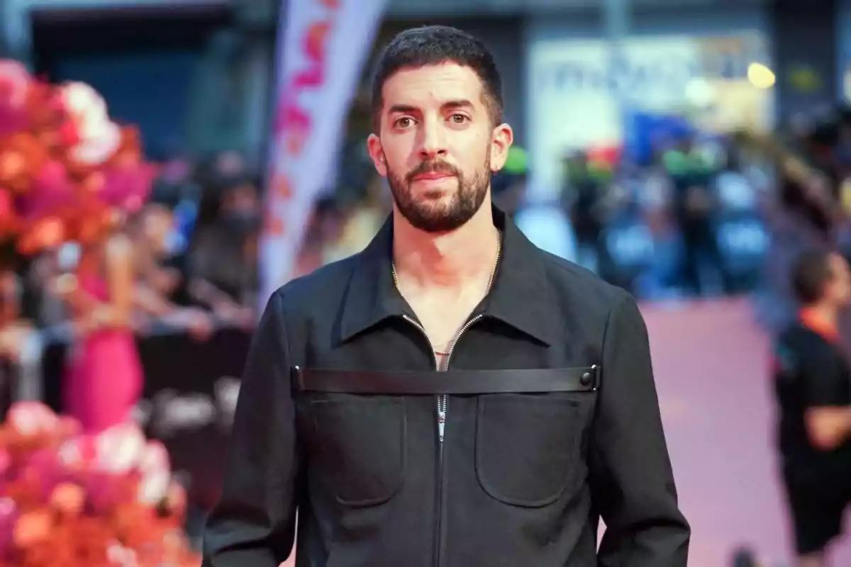 Hombre con barba y cabello corto vestido con chaqueta negra en una alfombra roja con fondo desenfocado de personas y flores rosas Hombre con barba y cabello corto vestido con chaqueta negra en una alfombra roja con fondo desenfocado de personas y flores rosas