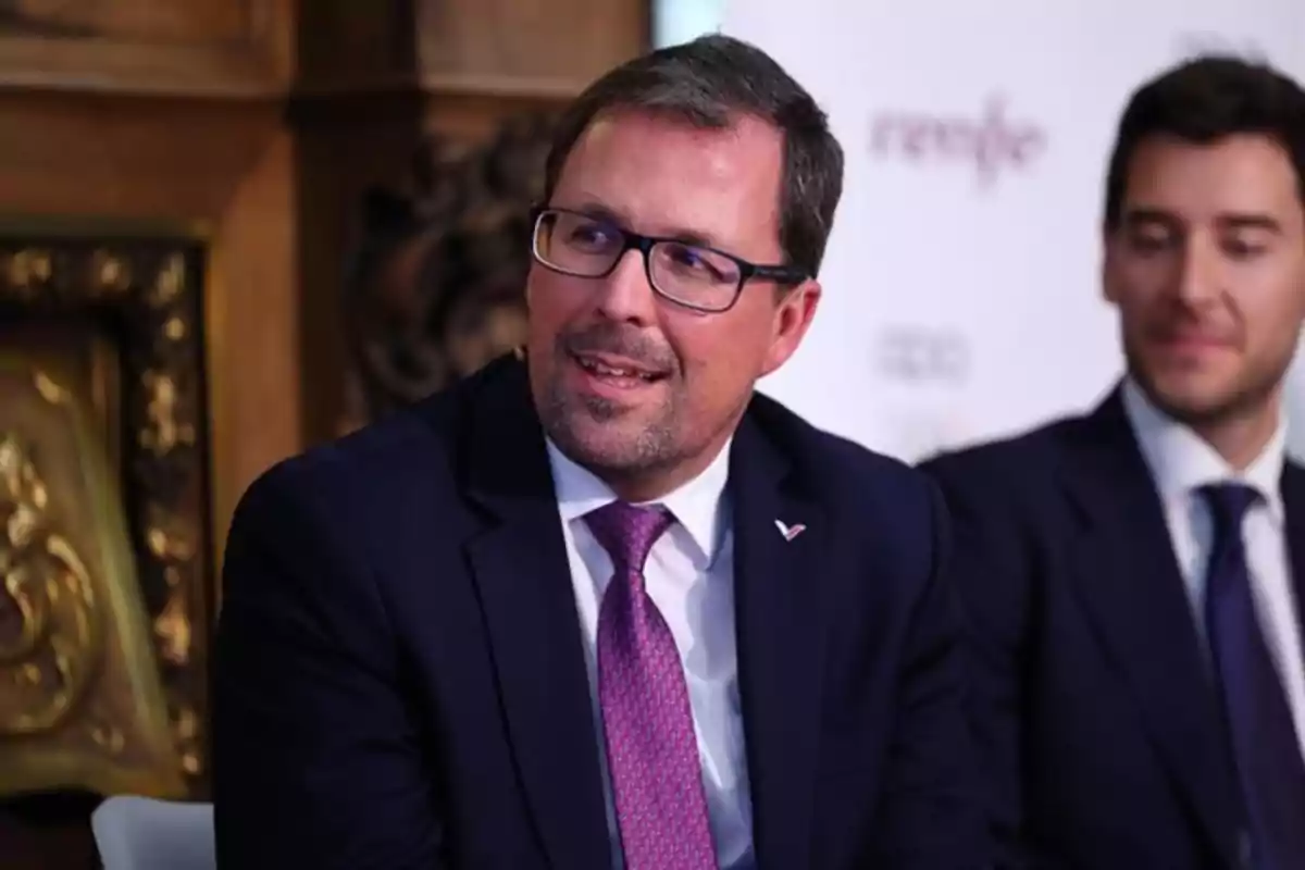 Hombre con gafas y traje oscuro sonríe mientras está sentado junto a otra persona en un evento formal Hombre con gafas y traje oscuro sonríe mientras está sentado junto a otra persona en un evento formal