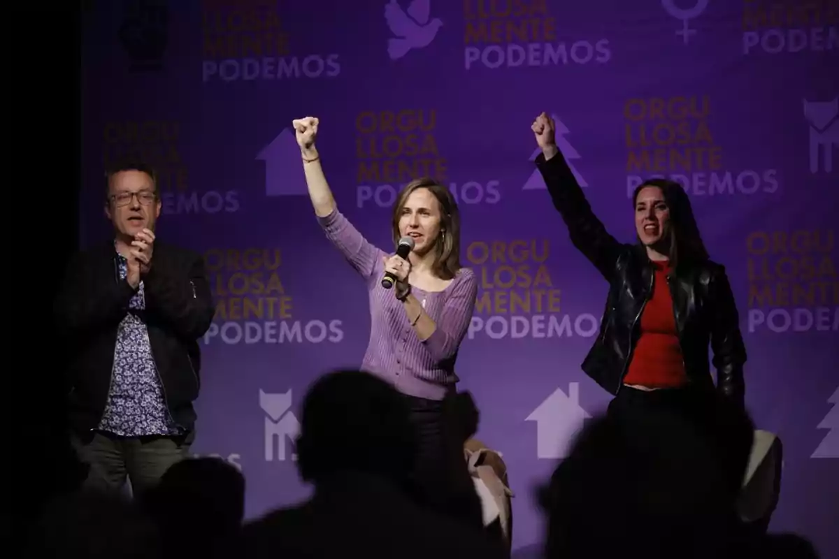 Tres personas en un escenario con fondo morado de Podemos, dos de ellas levantan el puño y una sostiene un micrófono, mientras el público observa Tres personas en un escenario con fondo morado de Podemos, dos de ellas levantan el puño y una sostiene un micrófono, mientras el público observa