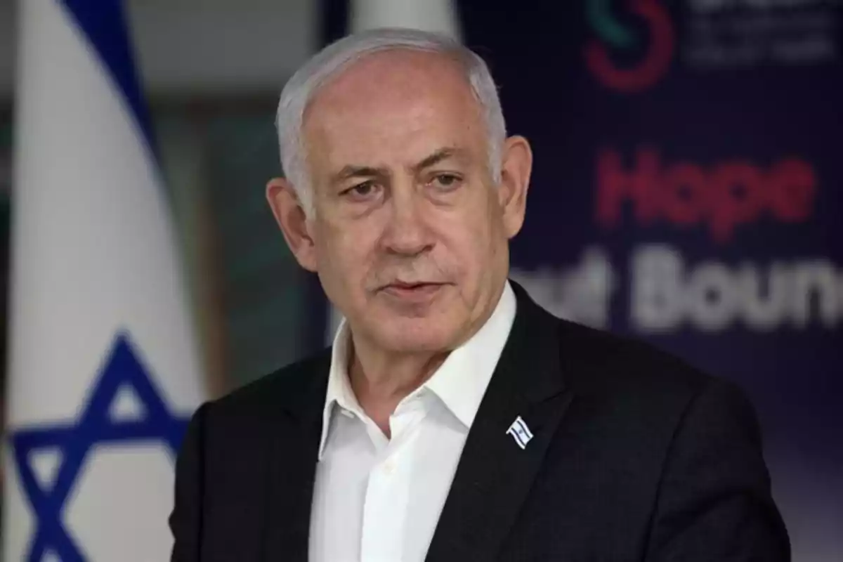 Hombre de cabello canoso con traje oscuro y camisa blanca, de pie frente a una bandera de Israel y un fondo con letras desenfocadas Hombre de cabello canoso con traje oscuro y camisa blanca, de pie frente a una bandera de Israel y un fondo con letras desenfocadas