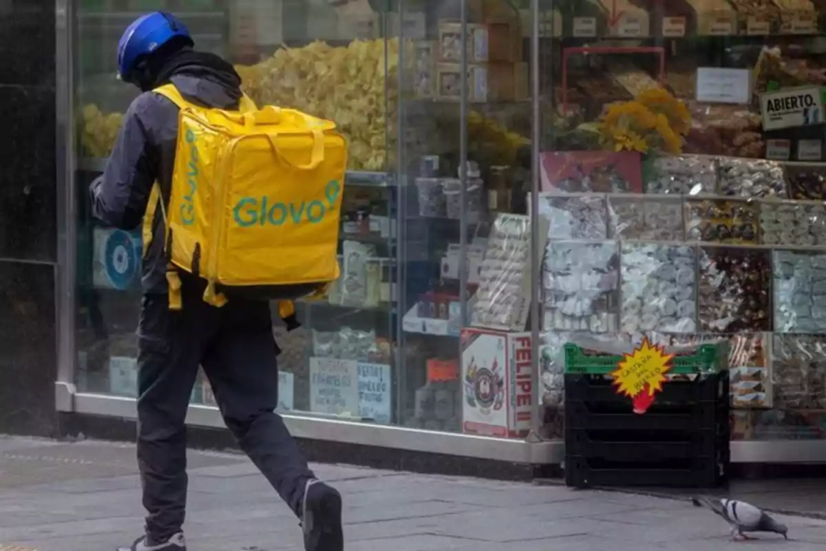 Repartidor de Glovo con mochila amarilla caminando frente a una tienda de dulces y frutos secos Repartidor de Glovo con mochila amarilla caminando frente a una tienda de dulces y frutos secos