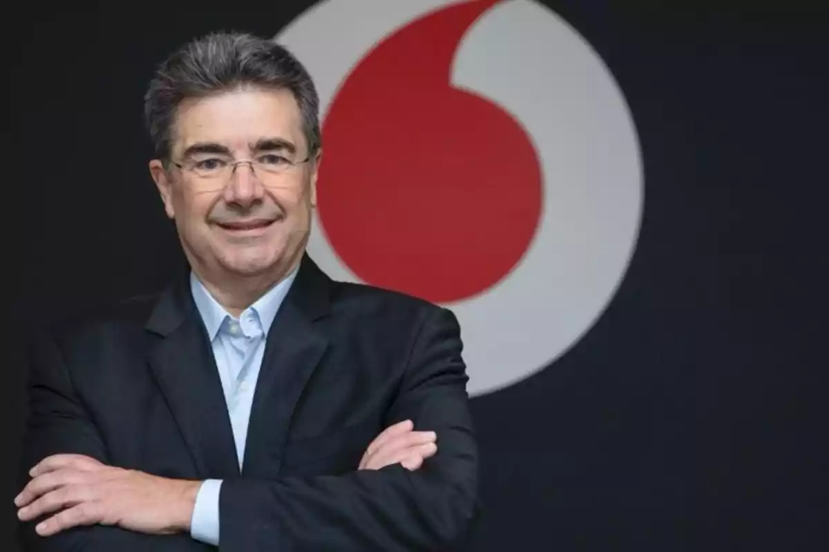 Hombre de cabello canoso y gafas con traje oscuro y camisa azul claro sonriendo frente a un logotipo circular rojo y blanco sobre fondo negro