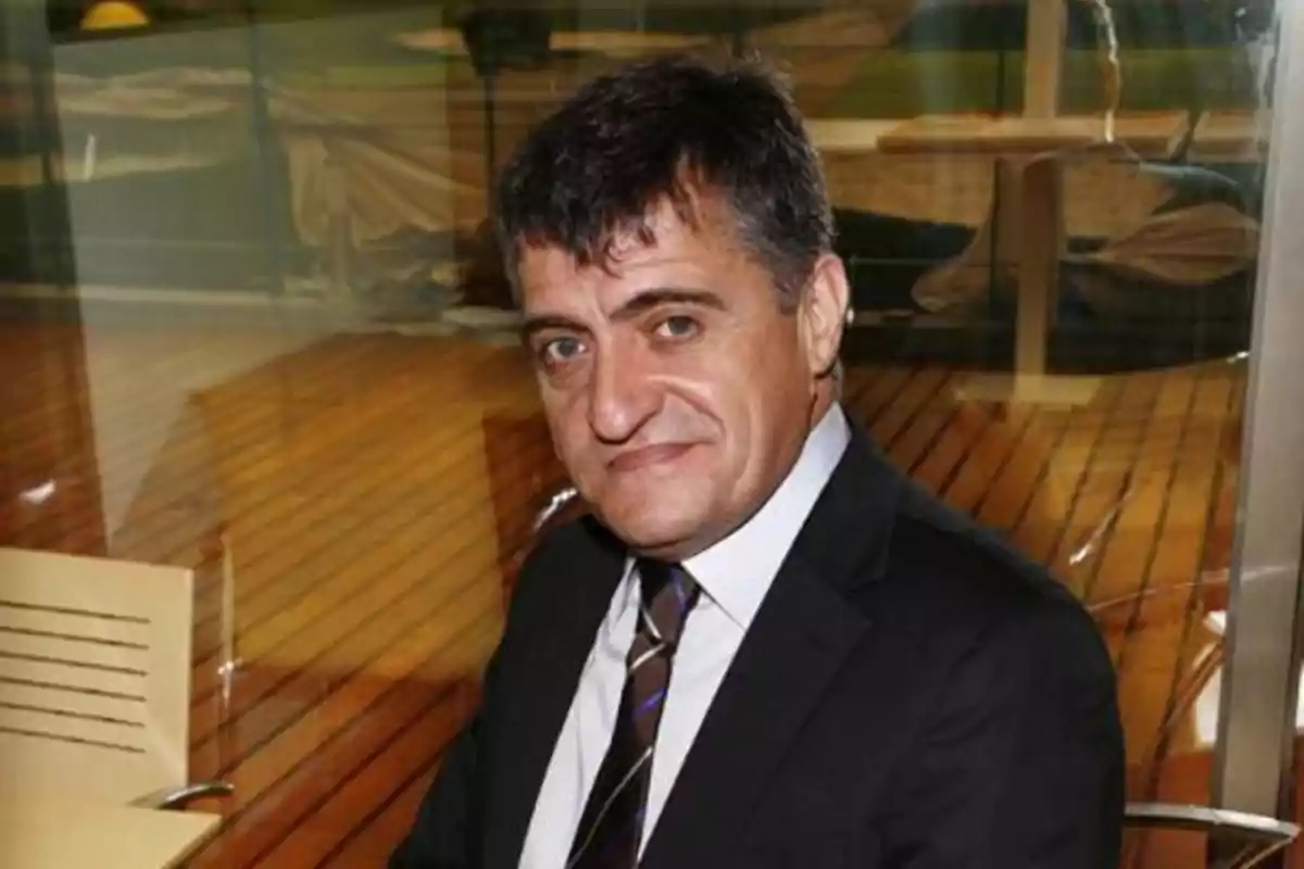 Hombre de cabello oscuro con traje y corbata sentado en una silla en un ambiente interior con piso de madera Hombre de cabello oscuro con traje y corbata sentado en una silla en un ambiente interior con piso de madera